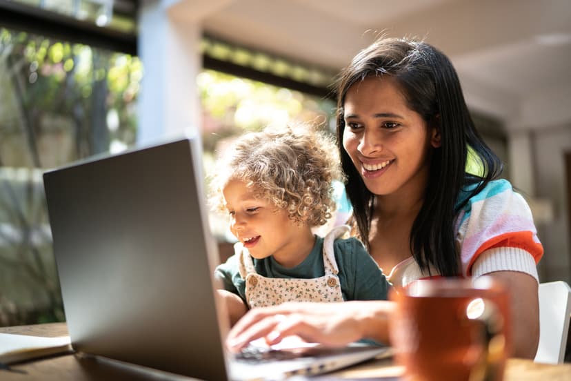 A mother using a laptop while holding her daughter at home
